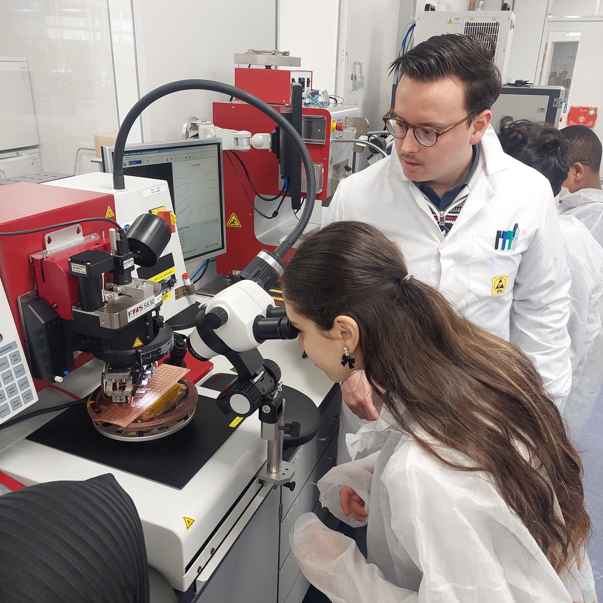 Child in labcoat looking through microscope under supervision of a scientist